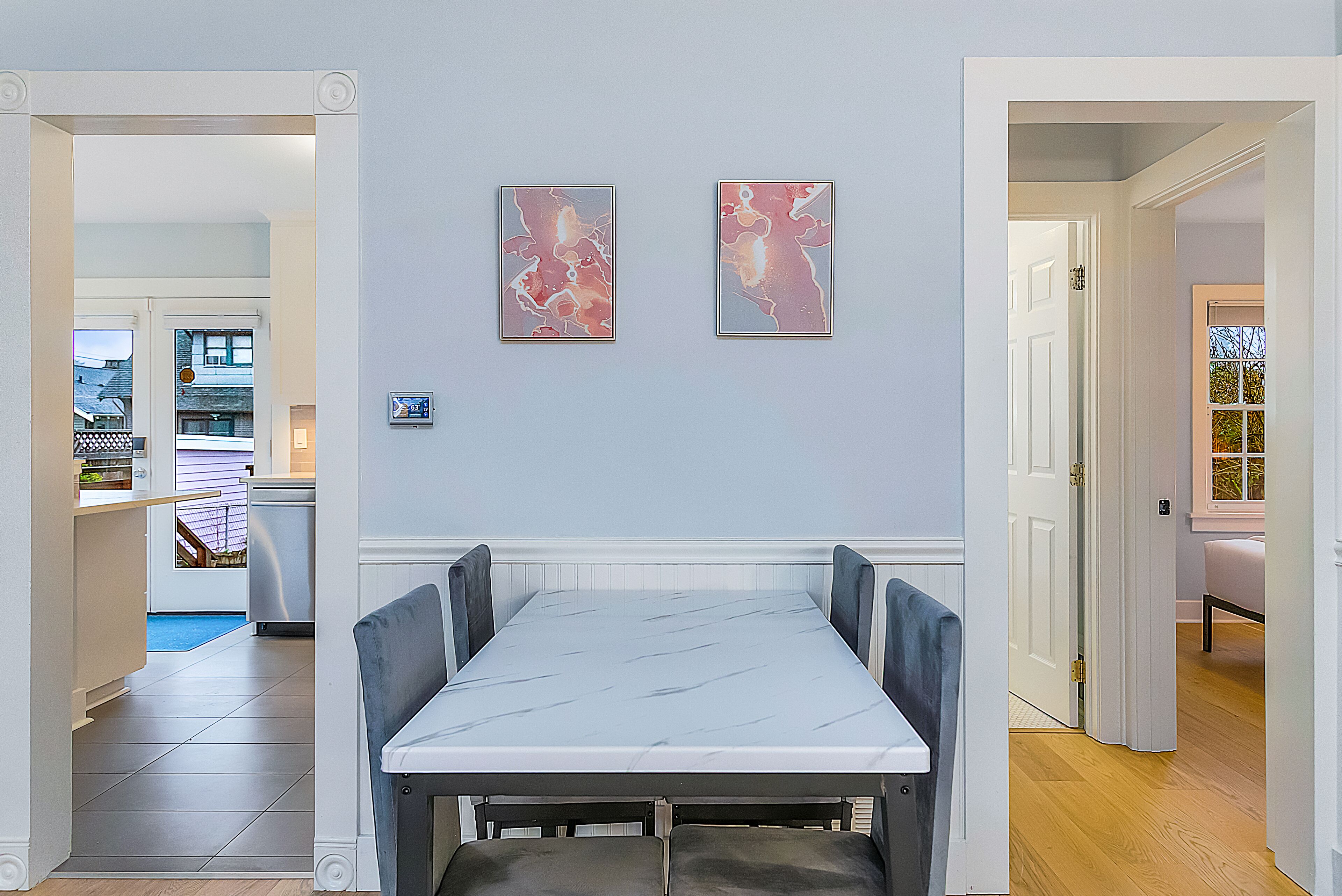 A dining area with a marble table and chairs, two framed artworks on the wall, and a view of the kitchen and living area in the background.