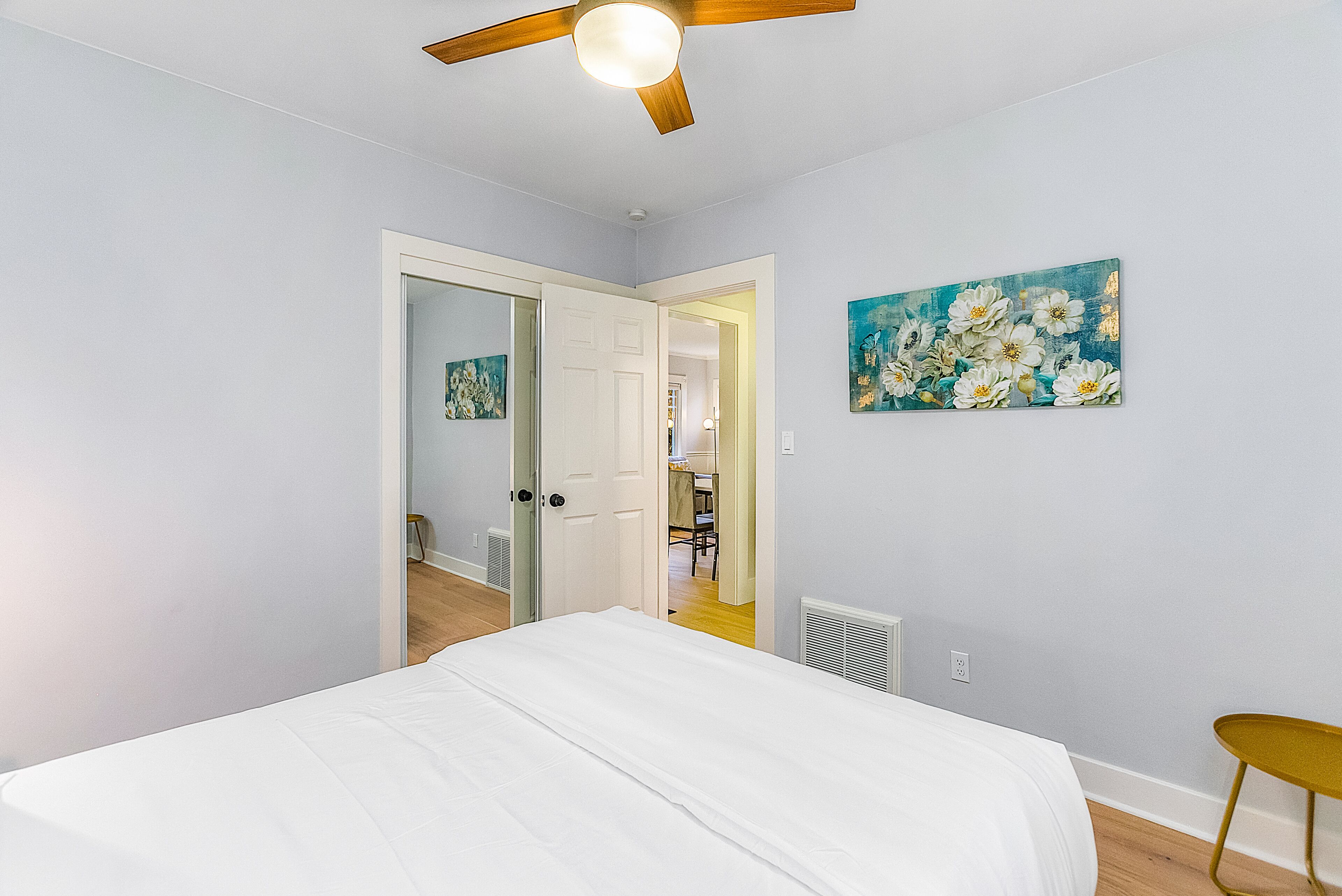 A bedroom with a bed, a ceiling fan, a painting on the wall, and a door leading to another room.
