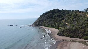 A coastal landscape with a beach, rocky shoreline, and a forested hill. - Oceanfront Condo with stunning pool and oversized balcony. (Jacó)