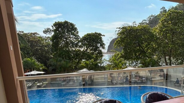 A pool with clear blue water, surrounded by lounge chairs and umbrellas, with a view of the ocean and trees in the background. - Oceanfront Condo with stunning pool and oversized balcony. (Jacó)