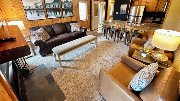A cozy living room with a brown leather sofa, a bench, and a coffee table. There is a kitchen area with a refrigerator, microwave, and bar stools. - 8 Powderidge, Snowshoe, WV - Slopeside Sanctuary (Snowshoe)