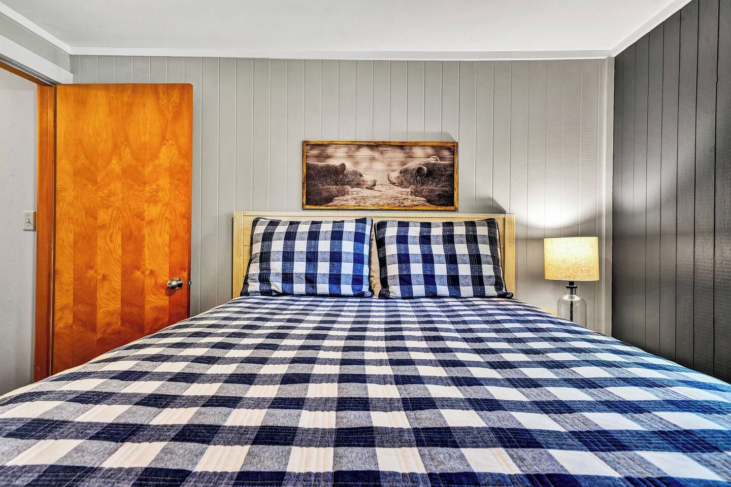 Hotellihuone, jossa on ruutukuviollinen peitto sängyllä, puinen ovi, seinällä oleva kehystetty kuva ja yöpöytälamppu.