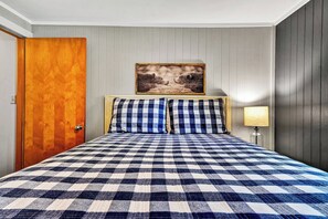 A hotel room with a checkered bedspread, a wooden door, a framed picture on the wall, and a bedside lamp. - 8 Powderidge, Snowshoe, WV - Slopeside Sanctuary (Snowshoe)