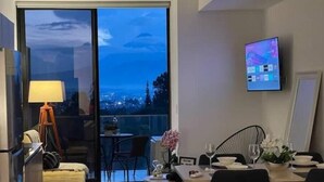 Una sala de estar moderna con comedor, televisor montado en la pared y un ventanal con vista a la ciudad.