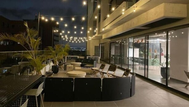 Un área de estar al aire libre con un sofá negro curvo, una mesa redonda y luces colgantes.