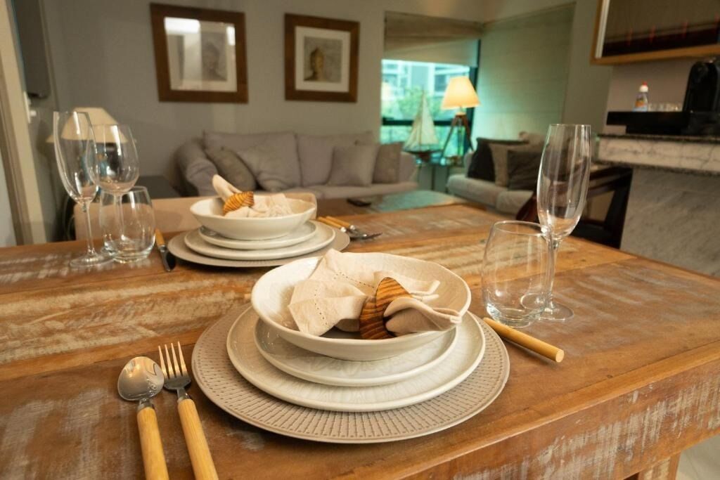 Una mesa de comedor dispuesta con platos, cuencos y cubiertos blancos, una mesa de madera y una vista de una sala de estar al fondo.