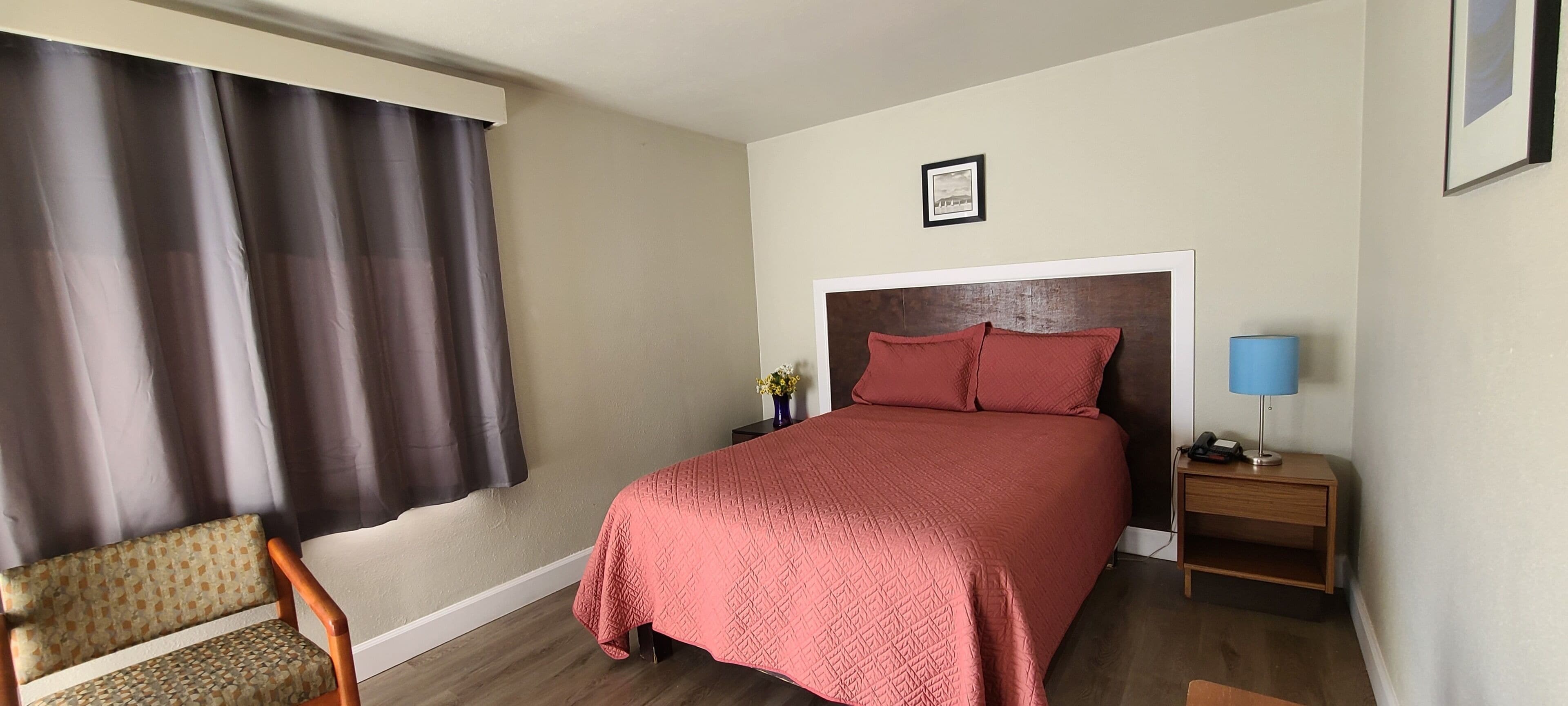 A bedroom with a bed, a nightstand, a chair, a window with curtains, and a framed picture on the wall.