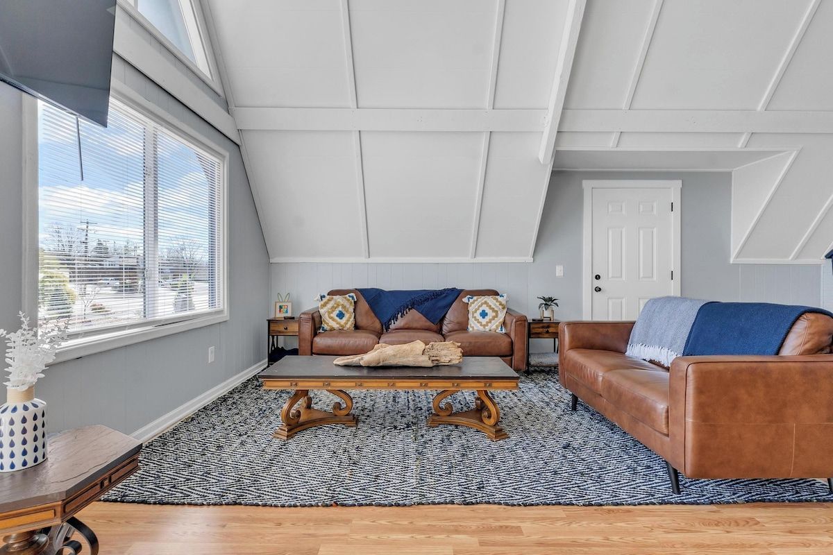 A loft-style room with a sloped ceiling, a brown leather sofa, a wooden coffee table, and a patterned rug.