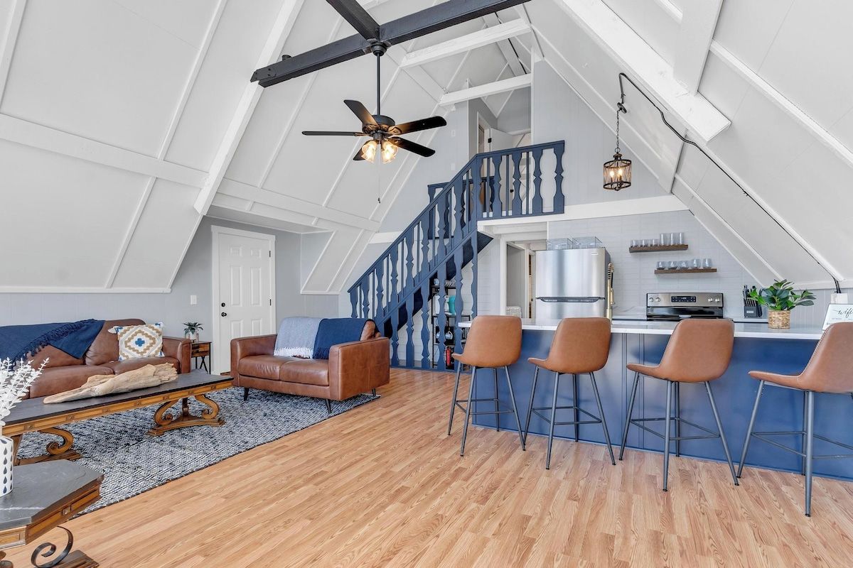 Un loft amplio con techo alto, área de cocina, zona de estar con sofá y sillas, y una escalera.