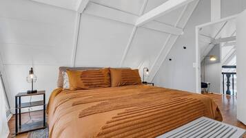 Un dormitorio con techo inclinado, una cama con colcha estampada, una mesita de noche con lámpara y una pequeña mesa frente a la cama.