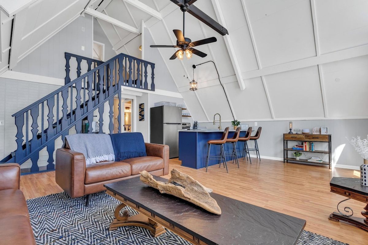 A spacious living area with a high ceiling, a staircase, a kitchen area, and a seating area with a sofa and coffee table.