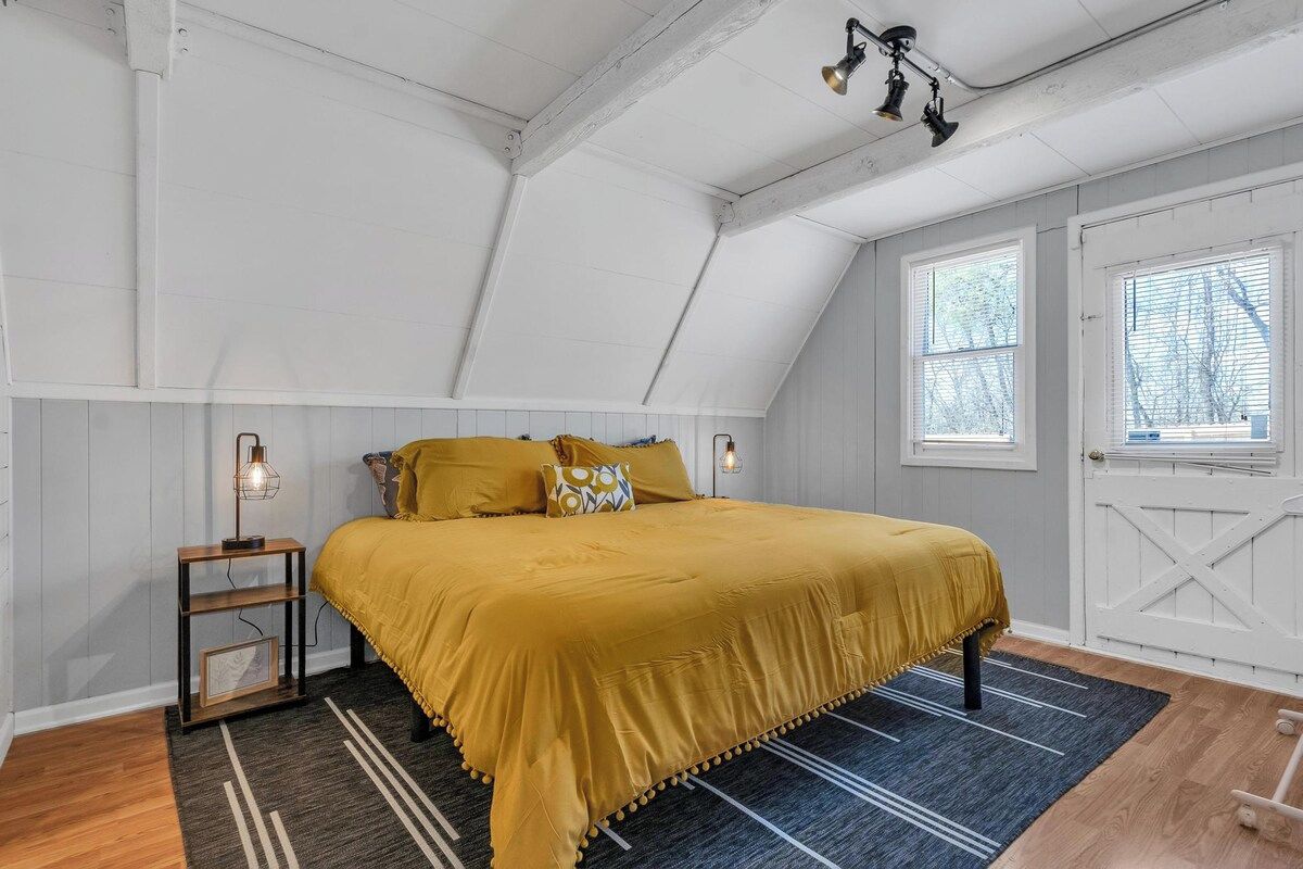 A bedroom with a sloped ceiling, a bed with a yellow bedspread, a nightstand with a lamp, and a window with blinds.
