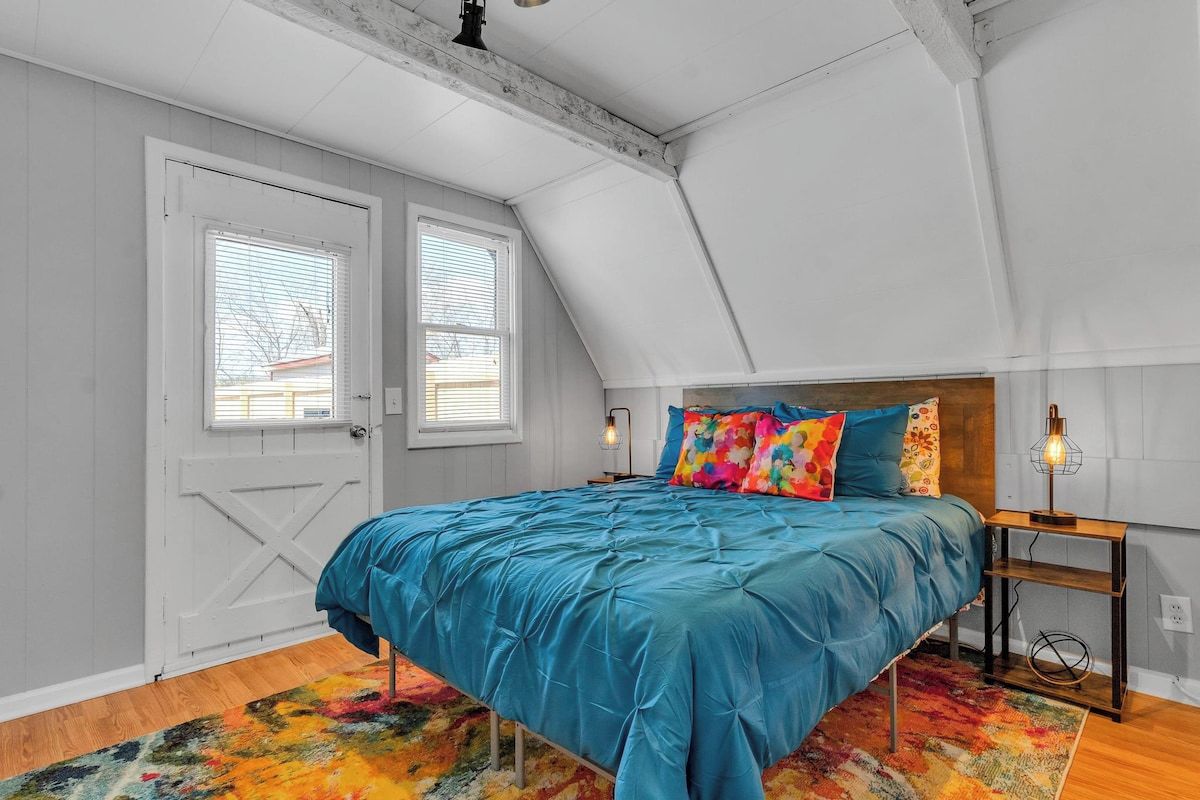 A bedroom with a sloped ceiling, a bed with a teal bedspread, colorful pillows, a multicolored rug, and a small bedside table with a lamp.