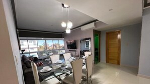 A modern dining area with a glass table and chairs, a television mounted on the wall, and a view of the outdoors through large windows. - Luxury & Secure-Santo Domingo Este-Close & Safe-Weekly Discounts (Santo Domingo Este)