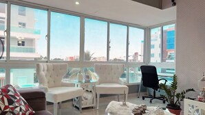 A modern living room with a sofa, two white armchairs, a black office chair, and a glass coffee table with decorative items. Large windows offer a view of the cityscape. - Luxury & Secure-Santo Domingo Este-Close & Safe-Weekly Discounts (Santo Domingo Este)