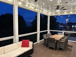 An outdoor deck with a wooden table and chairs, a white sofa with a red cushion, and string lights. - 1 min. golf cart ride to the beach! Family-sized house w/ deck & fenced backyard (Panama City Beach)