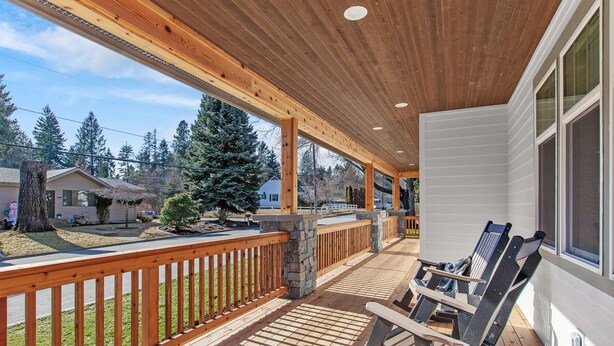 A covered porch with wooden railings and a stone wall, featuring two reclining chairs and a view of a residential area with houses and trees. - Tucked Away Paradise (Coeur d'Alene)