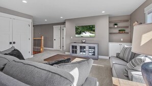 A spacious living room with grey sofas, a wooden coffee table, and a TV mounted on the wall. There are white cabinets below the TV and a staircase leading to another room. - Tucked Away Paradise (Coeur d'Alene)
