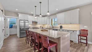 A spacious kitchen with white cabinets, a large island with bar stools, stainless steel appliances, and pendant lighting. - Tucked Away Paradise (Coeur d'Alene)