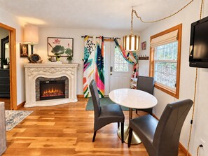 A living room with a fireplace, a round table with chairs, a television mounted on the wall, and a colorful curtain. - HOT TUB! Large deck, BBQ grill, fire pit & Arkansas Sunsets! (Eureka Springs)