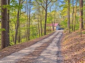A rural road leading to a single-story house with a red roof, surrounded by tall trees and a clear sky.