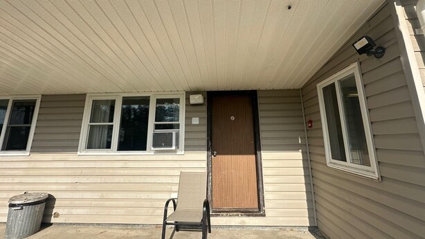 A building with a covered entrance, a wooden door, and a window with a view of trees. - Pine Ridge Motel (Timmins)