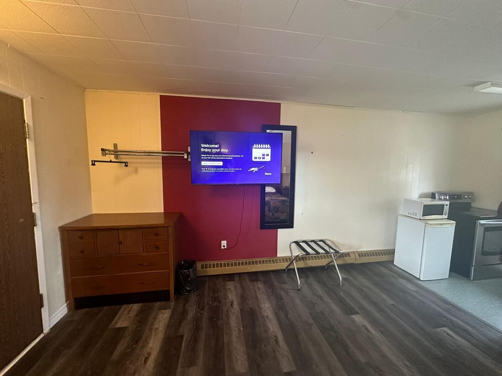 A hotel room with a wooden dresser, a flat-screen TV mounted on the wall, a small kitchenette with a microwave and toaster, and a radiator on the floor.