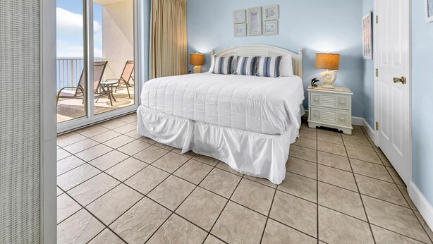 A bedroom with a large bed, a nightstand, a lamp, and a view of the balcony. - Ocean Ritz 1902 (Panama City Beach)