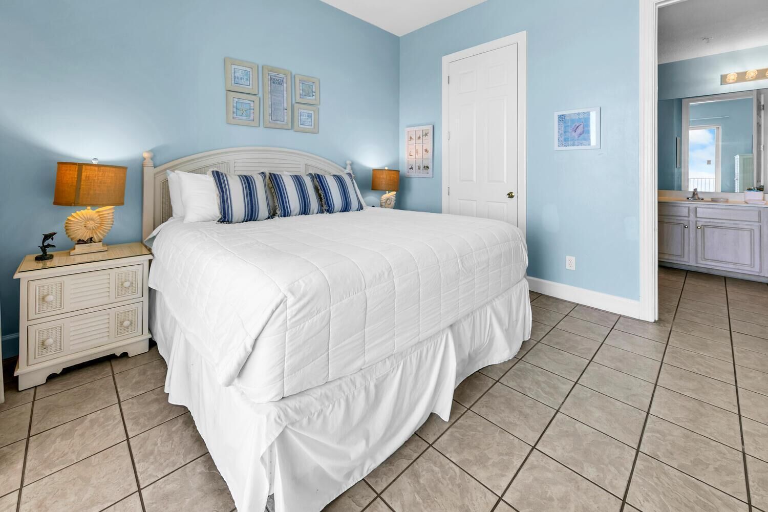 A bedroom with a large bed, two bedside tables with lamps, a door, and a bathroom visible in the background.