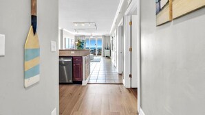 A hallway with wooden flooring, a kitchen area with a counter and cabinets, and a view to a balcony. - Tropic Winds 2308 (Panama City Beach)