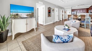 A spacious living room with a large flat-screen TV, a comfortable seating area, a kitchen with wooden cabinets, and a dining area with wicker furniture. - Tropic Winds 2308 (Panama City Beach)