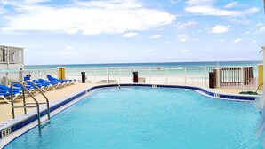 A pool with a view of the ocean, surrounded by blue lounge chairs and a white fence. - Tropic Winds 2308 (Panama City Beach)