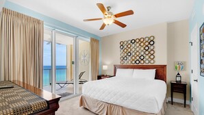 A hotel room with a large bed, a ceiling fan, a view of the ocean, and a wall decorated with circular mirrors. - Tropic Winds 1104 (Panama City Beach)