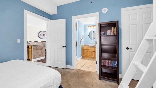 A bedroom with a bed, a bookshelf, and a bathroom visible through an open door. - Tropic Winds 1104 (Panama City Beach)