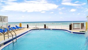 A pool with a view of the ocean, surrounded by blue lounge chairs and a white fence. - Tropic Winds 1104 (Panama City Beach)