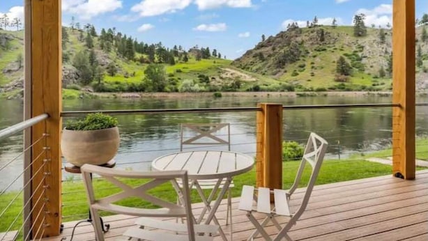 Una terraza de madera con una mesa redonda y sillas, con vistas a un lago tranquilo y colinas verdes.