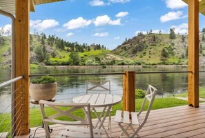A wooden deck with a round table and chairs, overlooking a calm lake and green hills. - Private Suite on the Missouri River (Cascade)