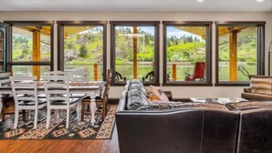 Una habitación con zona de comedor, sofá y ventanas amplias con vistas a un paisaje verde.