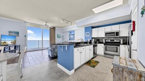 A modern kitchen with white cabinets, a blue island, and a large window overlooking the beach.