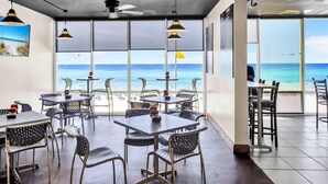 Un restaurant avec des tables et des chaises, de grandes fenêtres donnant sur la plage et des ventilateurs de plafond.