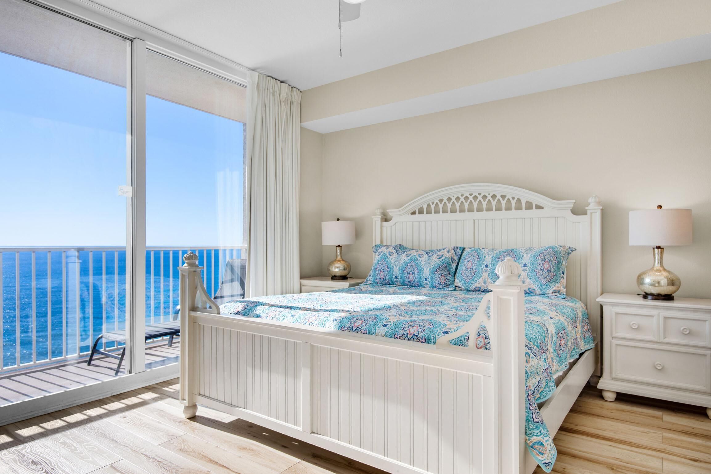 A bedroom with a balcony, a bed with patterned bedding, two bedside lamps, and a view of the ocean.