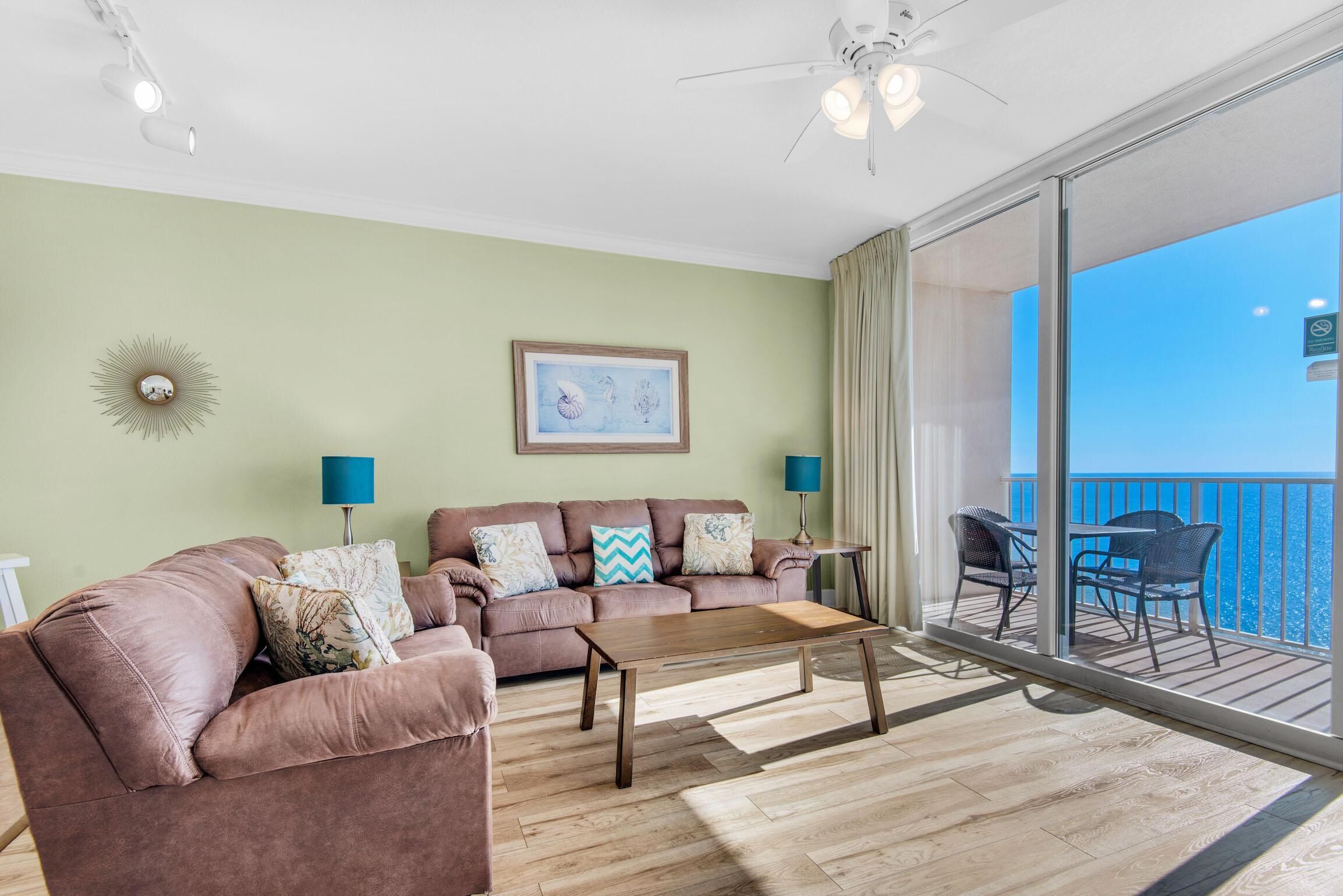 A living room with a sofa, coffee table, and two lamps. There is a balcony with chairs and a view of the sea.