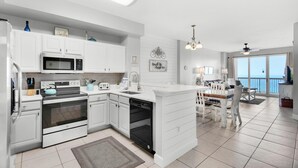 Una cocina moderna con armarios blancos, horno integrado, microondas y lavavajillas. Hay un comedor con una mesa y sillas blancas, y vistas al mar a través de amplios ventanales.