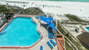 Una piscina con agua azul, rodeada de sillas de descanso y sombrillas, con una playa y el océano al fondo.