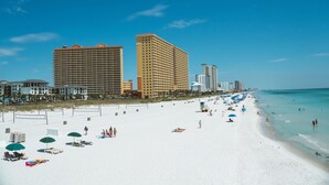 Ein Strand mit weißem Sand, Sonnenschirmen und Menschen. Im Hintergrund sind hohe Gebäude zu sehen.