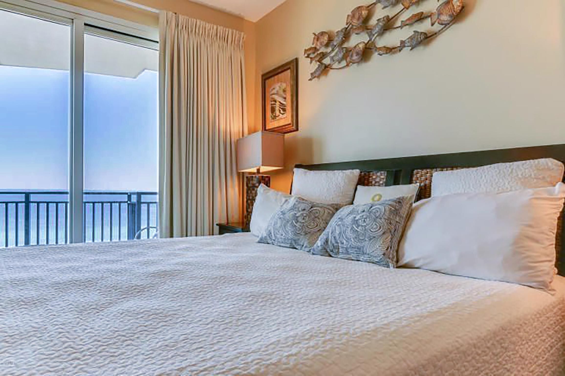 A hotel room with a large bed, a view of the sky through the window, and a decorative wall piece.