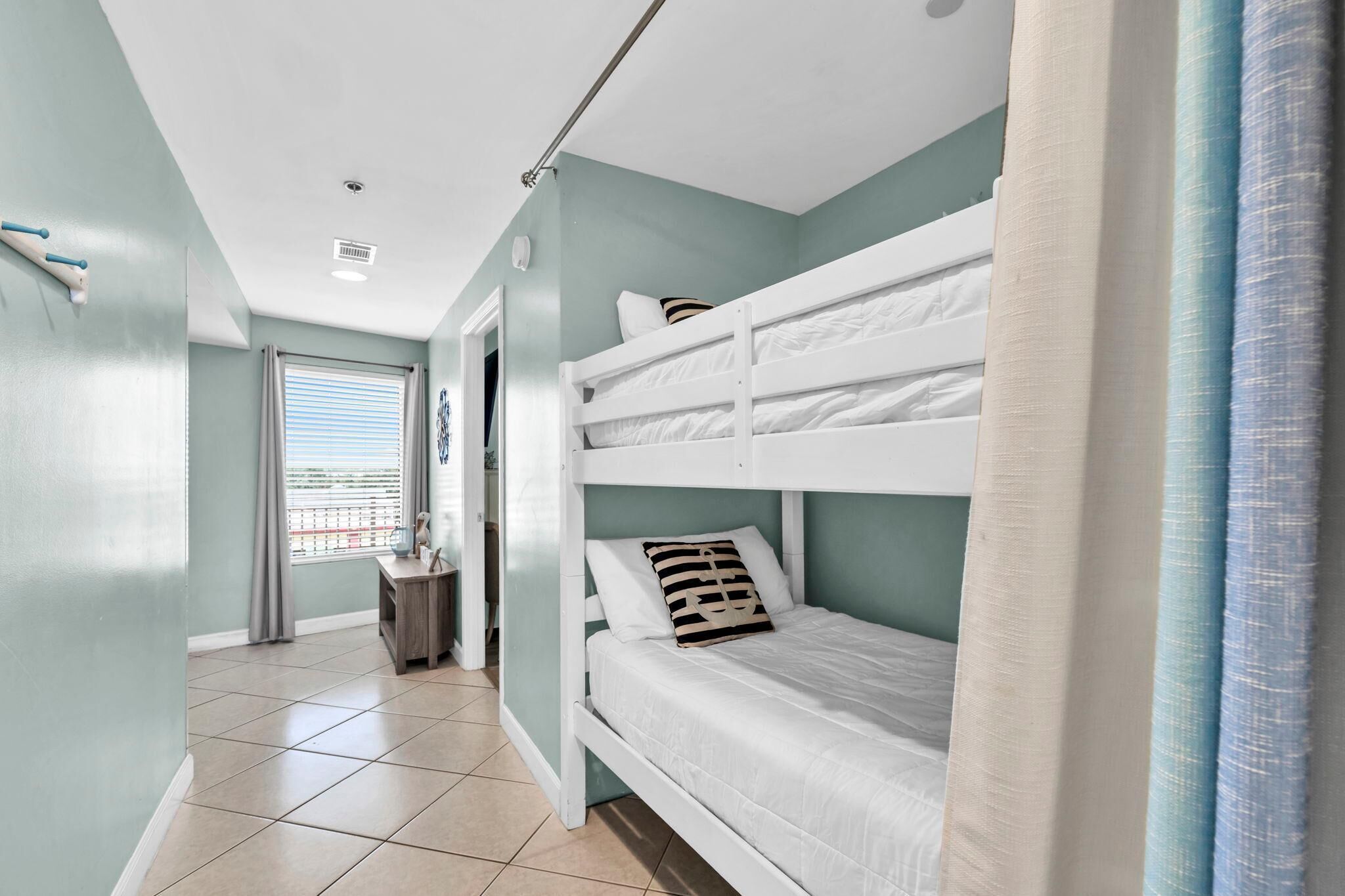 A bunk bed room with a view of the sea through a window, a small table, and a door leading to another room.
