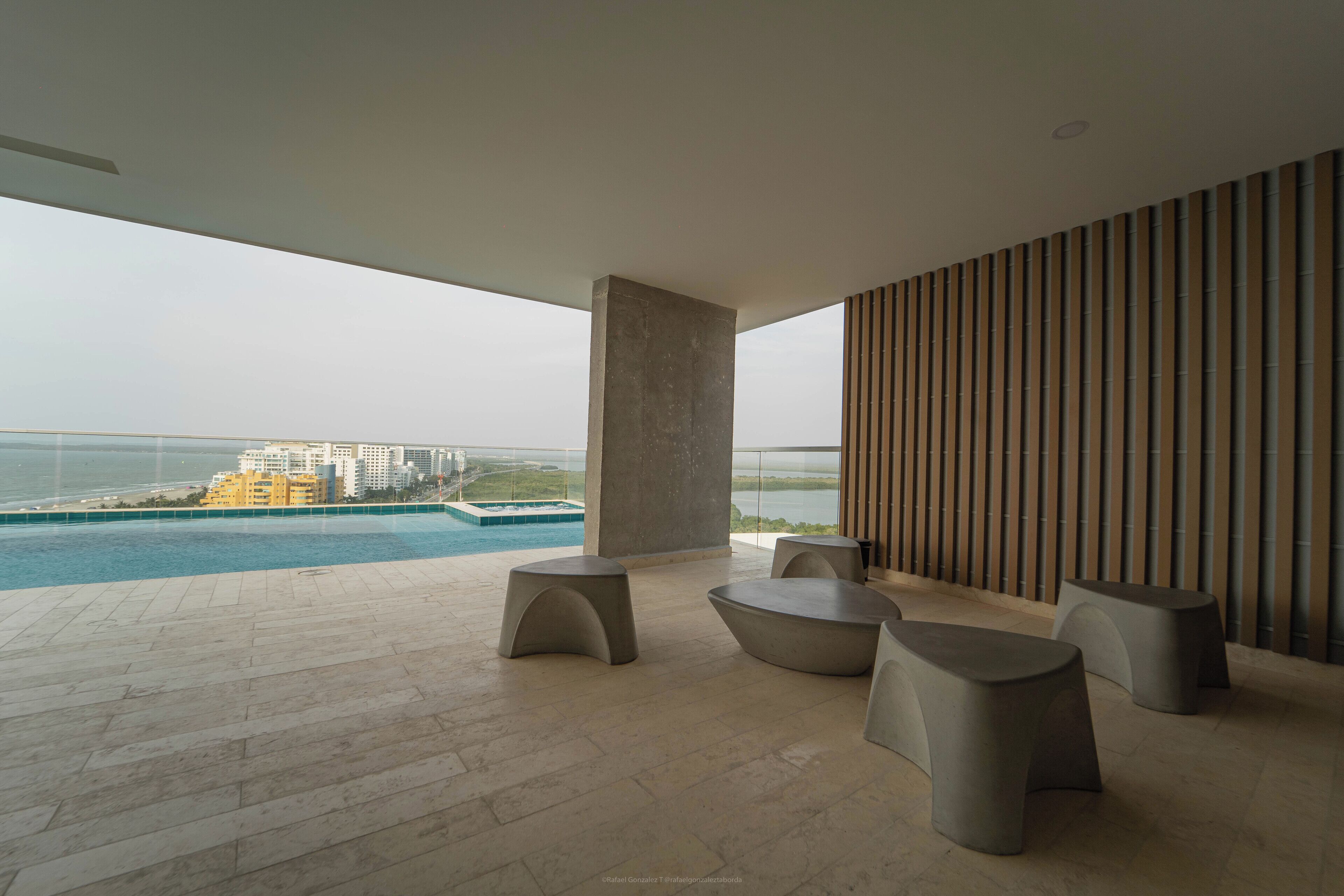 Un área lounge moderna al aire libre con piscina, asientos y vista al océano y edificios.