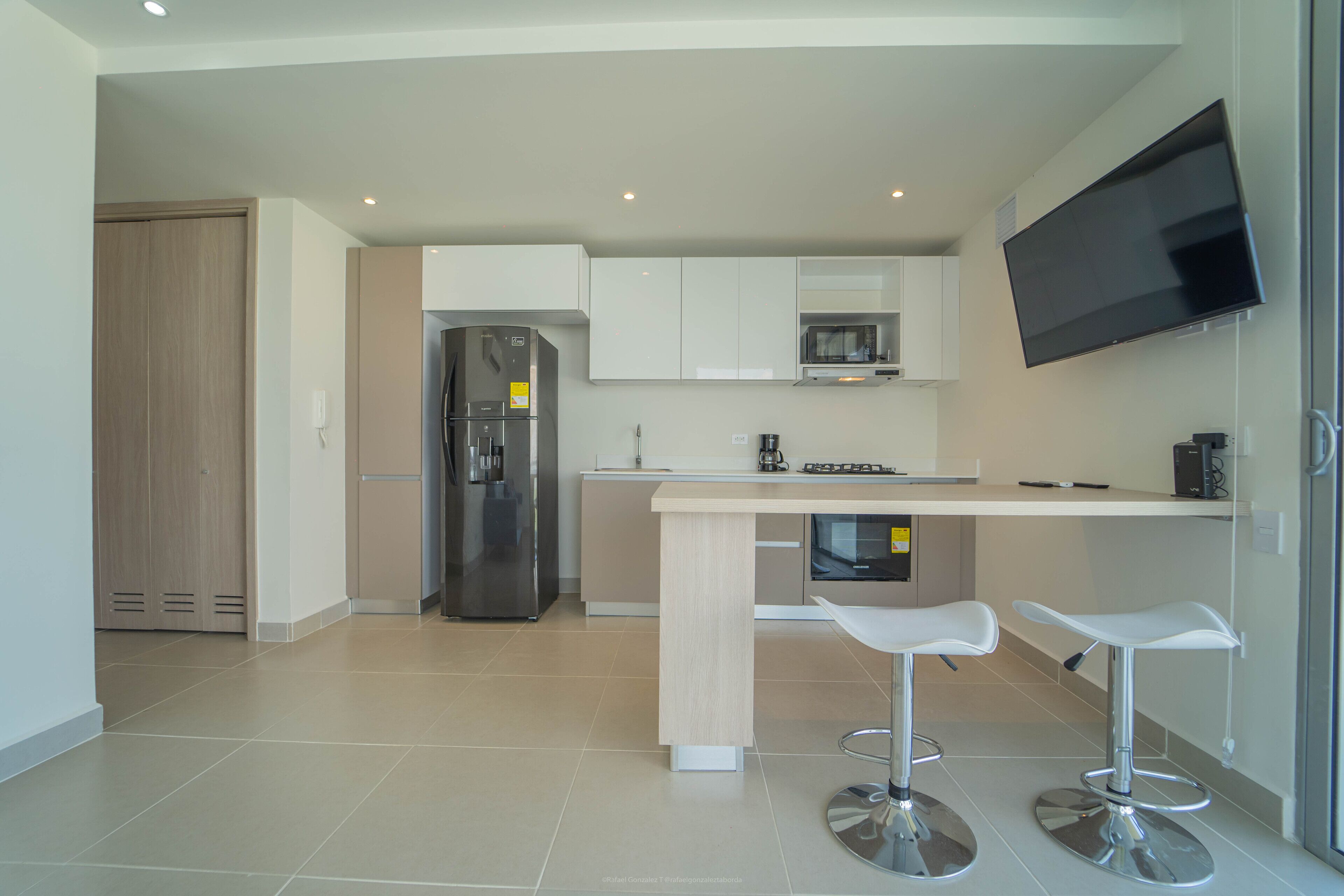 Una cocina moderna con refrigerador integrado, microondas y cafetera. Hay dos taburetes blancos y un televisor de pantalla plana montado en la pared.
