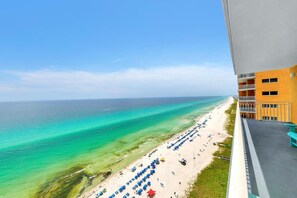 Blick auf einen Strand vom Balkon aus, mit Blick auf einen Sandstrand, blaue Sonnenschirme und ein klares, blaues Meer.